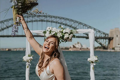 Smiling bride holds up a bouquet with wedding ach in background